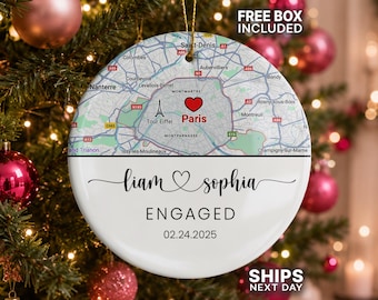 May include: A round white ceramic ornament featuring a map of Paris with a red heart icon over the city center. Below the map, the text reads Liam and Sophia, ENGAGED, and the date 02.24.2025 in black lettering. A gold string is attached to the top.