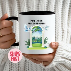 May include: White ceramic mug with a black interior and handle. The mug features an illustration of an archway with doves, trees, and the text "POPE LEO XIV: PEACE & PROGRESS".