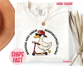 May include: Ivory-colored t-shirt featuring a cartoon duck wearing sunglasses and a red cap, riding a red scooter. The text "BEHAVIOR GOES WHERE REINFORCEMENT FLOWS" is printed around the duck. The shirt also has the text "MORE COLORS" and "SHIPS FAST".