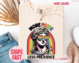 Könnte beinhalten: Elfenbeinfarbenes T-Shirt mit einem Aufdruck einer Statue mit herzförmiger Regenbogen-Sonnenbrille, einem Regenbogen und dem Text "MORE PRIDE LESS PREJUDICE". Das T-Shirt ist gefaltet, mit einer Halskette und Jeans sichtbar.