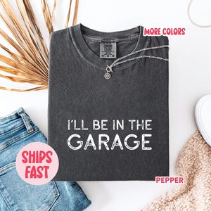 Könnte beinhalten: Dunkelgraues T-Shirt mit dem weißen Text "I'll be in the Garage". Das Bild zeigt blaue Jeansshorts, weiße Turnschuhe und eine silberne Halskette. Die Wörter "More Colors" und "Ships Fast" sind ebenfalls sichtbar.