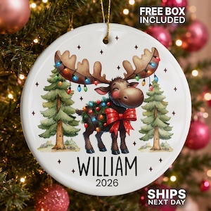 Op de afbeelding: Een ronde witte keramische kerstornament met een cartoon-eland met een gewei vol gekleurde kerstverlichting, een rode strik en twee dennenbomen. De naam WILLIAM en het jaartal 2026 staan in zwarte letters onderaan de afbeelding.