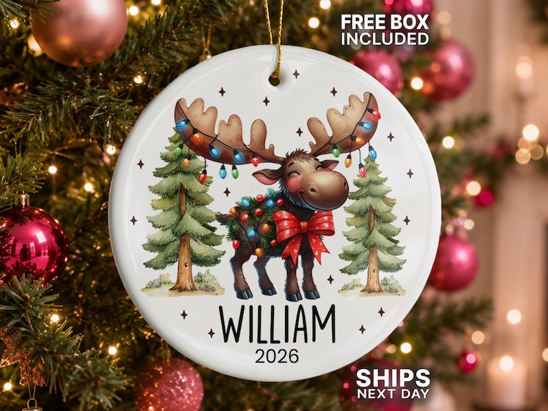 Op de afbeelding: Een ronde witte keramische kerstornament met een cartoon-eland met een gewei vol gekleurde kerstverlichting, een rode strik en twee dennenbomen. De naam WILLIAM en het jaartal 2026 staan in zwarte letters onderaan de afbeelding.