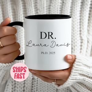 Könnte beinhalten: Eine weiße Kaffeetasse mit schwarzem Rand und Henkel. Die Tasse trägt einen schwarzen Schriftzug mit der Aufschrift "DR. Laura Davis Ph.D. 2025".
