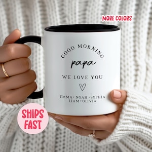Op de afbeelding: Witte keramische mok met een zwart handvat en interieur. De mok heeft de tekst "GOOD MORNING papa WE LOVE YOU" met een hartsymbool en namen. Een roze sticker met "MORE COLORS" is zichtbaar.