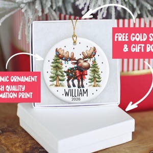 Op de afbeelding: Een rond wit keramieken ornament met een cartoon-eland versierd met kleurrijke kerstverlichting en een rode strik, staand tussen twee dennenbomen. De naam WILLIAM en het jaartal 2026 zijn in zwarte letters onder de eland afgebeeld.