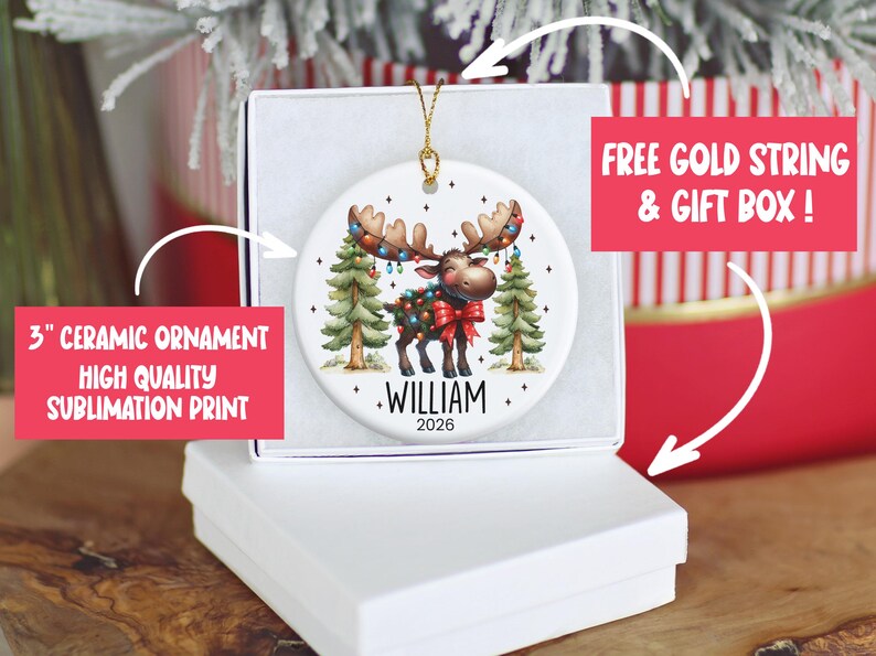 Op de afbeelding: Een rond wit keramieken ornament met een cartoon-eland versierd met kleurrijke kerstverlichting en een rode strik, staand tussen twee dennenbomen. De naam WILLIAM en het jaartal 2026 zijn in zwarte letters onder de eland afgebeeld.