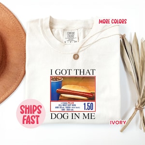 May include: Ivory-colored t-shirt with the text "I GOT THAT DOG IN ME" and a graphic of a hot dog and soda. The shirt also has the text "MORE COLORS" and "SHIPS FAST".