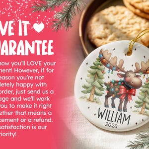 Op de afbeelding: Een rond wit keramieken ornament met een cartoon-eland met een gewei versierd met kleurrijke kerstverlichting, staand tussen twee dennenbomen. De naam WILLIAM en het jaartal 2026 staan in zwarte letters onder de illustratie van de eland.