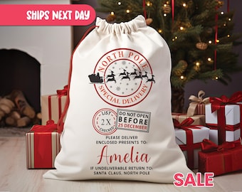 May include: A large white Christmas gift bag with a red drawstring, featuring a North Pole Special Delivery design. The bag includes the name "Amelia" and instructions for delivery and returns. Presents and a Christmas tree are in the background.