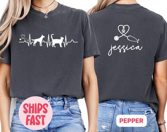 May include: Two dark gray t-shirts with a white graphic design. The front shirt has a heartbeat line with a dog and cat silhouette. The back shirt has a stethoscope with a heart and the name "jessica".