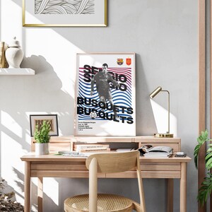 K&ouml;nnte beinhalten: Ein gerahmtes Poster mit einem Schwarzwei&szlig;bild eines Fu&szlig;ballspielers in einem blau-roten Trikot. Das Poster tr&auml;gt den Text "Sergio Busquets" in Schwarzwei&szlig;. Das Poster h&auml;ngt an einer Wand &uuml;ber einem Holztisch mit einer Lampe und einer Pflanze.