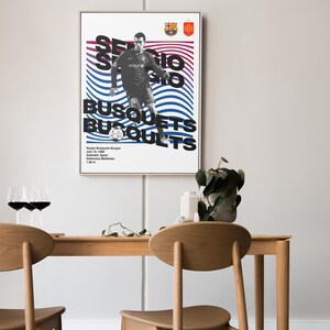K&ouml;nnte beinhalten: Ein schwarz-wei&szlig;es Poster mit einem grafischen Design eines Fu&szlig;ballspielers in einem blau-wei&szlig;en Trikot. Das Poster tr&auml;gt den Text "Sergio Busquets" in fetten schwarzen Buchstaben. Das Poster ist gerahmt und h&auml;ngt an einer wei&szlig;en Wand.