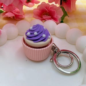 May include: A cupcake keyring featuring a light pink fluted base and a purple swirled frosting top decorated with small white dots. The cupcake is attached to a silver-toned metal split ring and a lobster claw clasp for secure attachment to items.