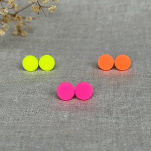 Könnte beinhalten: Eine Sammlung von drei Paar Ohrsteckern in leuchtenden Farben. Die Ohrringe sind in Neon-Gelb, leuchtendem Orange und Pink gehalten. Jedes Paar hat eine einfache, kreisrunde Form. Die Ohrringe liegen auf einem grauen Stoff.