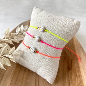 Könnte beinhalten: Drei Freundschaftsarmbänder mit neonrosa, neongelb und orangenen Schnüren. Jedes Armband hat einen weißen Smiley-Anhänger mit goldener Umrandung.