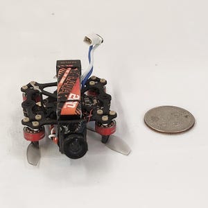 May include: A small black and red drone with propellers, a camera, and the text "LAVA 20". A quarter is next to the drone for size comparison. The drone has a white and blue wire attached.