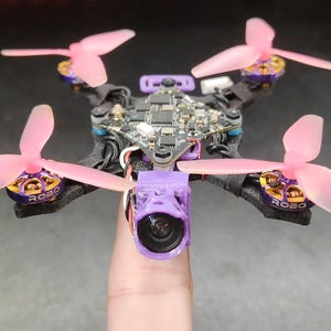 Peut inclure: Un petit drone quadricoptère doté d'un châssis en fibre de carbone noir, de quatre hélices tripales rose translucide et de moteurs dorés marqués ROBO. L'appareil comprend une carte électronique centrale et un support de caméra violet.