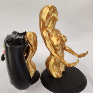 Könnte beinhalten: Eine goldfarbene, geometrisch geformte Figur eines Menschen mit ausgestreckten Armen, die einen schwarzen Videospiel-Controller hält. Die Figur steht auf einem schwarzen Sockel.