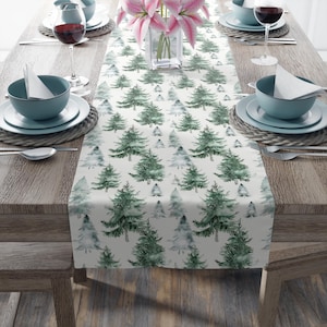 May include: A white table runner with a repeating pattern of green and gray watercolor pine trees. The runner is set on a wooden table, with place settings and a vase of pink flowers visible.
