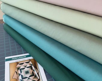 May include: A stack of five folded fabric bolts in a gradient of colors ranging from cream to dark forest green, accompanied by a quilt pattern booklet titled Percussion by Lisa Lund for Shadow Valley Quilts, featuring a beginner skill level.