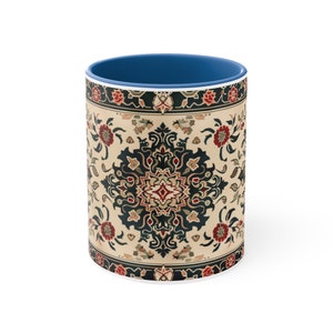 May include: A white ceramic mug with a blue interior. The exterior features a beige background with a detailed floral pattern in red, black, and dark blue. The design resembles a traditional rug.