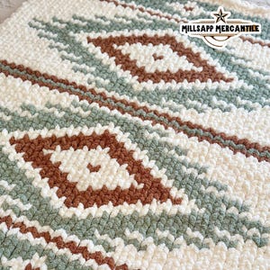 May include: A close-up of a soft, textured blanket with a geometric pattern. The blanket features a repeating design of white, sage green, and brown diamond shapes and stripes. The blanket is made of a thick, plush material.