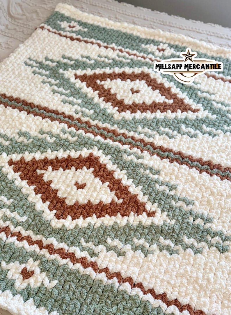 May include: A close-up of a soft, textured blanket with a geometric pattern. The blanket features a repeating design of white, sage green, and brown diamond shapes and stripes. The blanket is made of a thick, plush material.