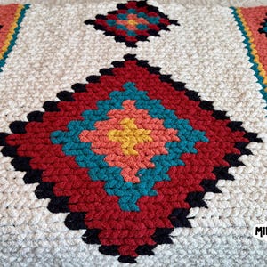 May include: A hand-knitted blanket with a geometric pattern in red, teal, coral, yellow, black, and white. The blanket has a diamond pattern in the center and along the edges. The blanket is made of a soft, chunky yarn.