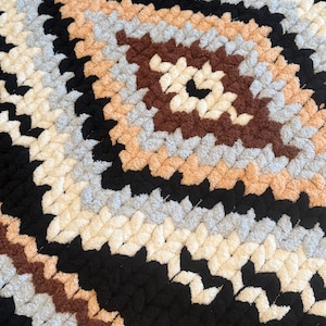 May include: A handmade, plush blanket with a geometric pattern. The blanket features a repeating diamond design in black, cream, brown, grey, and peach colours. The thick, textured yarn creates a cosy, inviting look.
