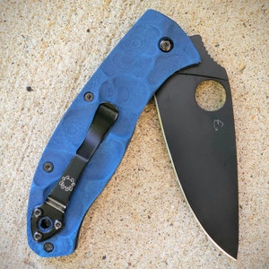 Può includere: Un coltello pieghevole blu con una lama nera. L'impugnatura presenta un motivo strutturato e una clip da tasca nera con un logo a ragno. La lama ha un ritaglio circolare e un piccolo segno.