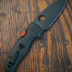 May include: Black folding knife with a textured handle and a black blade. The knife has a red thumb stud and a black pocket clip. The blade has a circular cutout and the text "Golden, Colorado U.S.A. Earth".