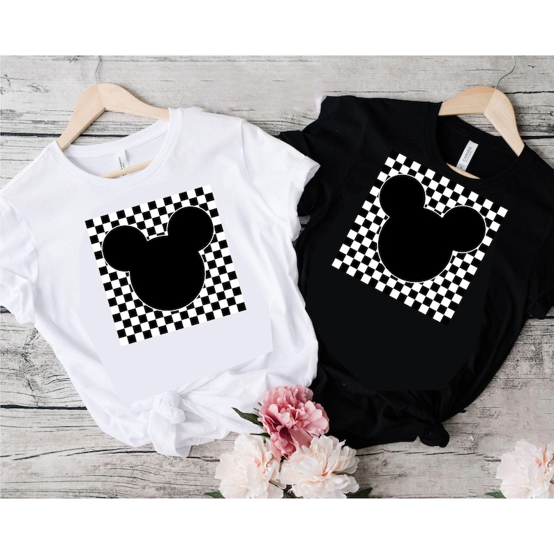 Puede incluir: Dos camisetas, una blanca y otra negra, cada una con un dise&ntilde;o de cabeza de Mickey Mouse negro sobre un fondo a cuadros. Las camisetas se muestran en perchas de madera sobre un fondo de madera clara, con flores rosas en primer plano.