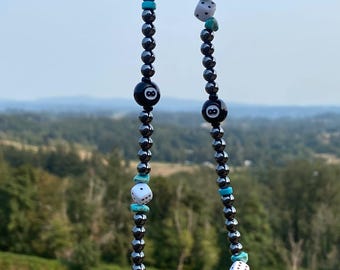 May include: A beaded necklace with black hematite beads, turquoise accents, and white dice and black 8-ball charms. The necklace is held up against a backdrop of a green field and a distant mountain range.
