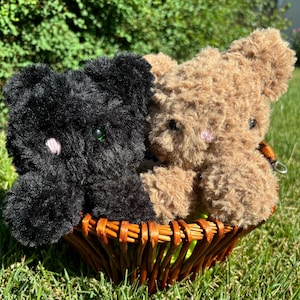 May include: Two plush teddy bears, one black and one brown, sitting in a wicker basket on a bed of green grass.