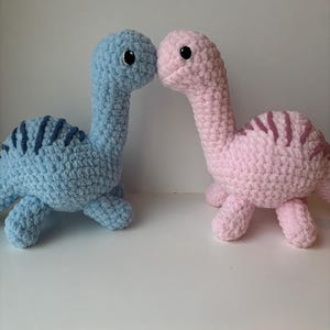 May include: Two crocheted dinosaur plushies, one blue and one pink, facing each other. The blue dinosaur has a dark blue stripe on its back, and the pink dinosaur has a dark purple stripe on its back.