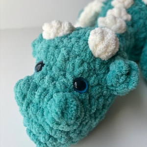 May include: A teal blue crocheted dragon plush toy with white yarn accents and black eyes.