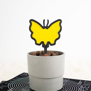 Peut inclure: Un piquet de plante en métal en forme de papillon jaune est placé dans un pot en béton gris rempli de billes d'argile brunes. Le pot est posé sur un livre noir intitulé "IRIDESCENT" et le texte "Holographics in design".