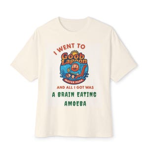 May include: Cream-colored t-shirt with the text "I WENT TO Goob Lagoon WATER PARK AND ALL I GOT WAS A BRAIN EATING AMOEBA." The graphic features a cartoon character in a water park setting. The shirt is a casual wear item.