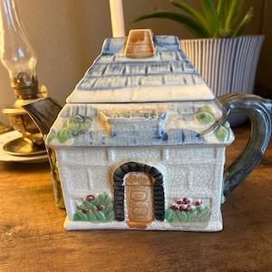 Può includere: Teiera in ceramica a forma di casa, con tetto blu, porta marrone e dettagli verdi. La teiera ha un manico e un beccuccio scuri, con finitura craquelé. Un oggetto da cucina unico, la teiera è un affascinante elemento decorativo.