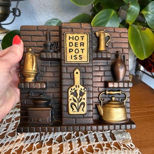 May include: A decorative wall hanging featuring a brick design with miniature kitchen items. The piece includes a kettle, teapot, and a cutting board. The text "HOT DER POT ISS" is displayed on a sign. The color scheme is brown and gold.