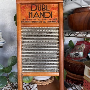 Op de afbeelding: Een vintage houten wasbord met een metalen schrobvlak. Het bovenste gedeelte bevat de woorden "DUBL HANDI" in vette letters, met extra tekst eronder. Het wasbord is gemaakt van hout en heeft een rechthoekige vorm.