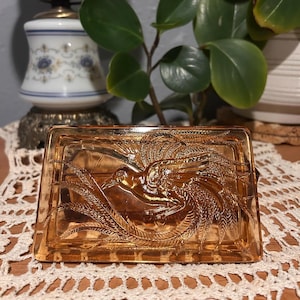 May include: A rectangular, amber-colored glass dish with a detailed embossed design of a bird and foliage. The dish is likely vintage, with a decorative and functional purpose. The glass has a warm, inviting tone, and the design is intricate.