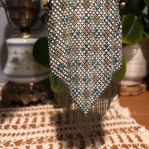 May include: A metal mesh purse featuring a geometric pattern in white, teal, and gold tones. The bag has a gold-toned metal frame at the top and a long, dangling fringe made of thin gold-toned metal chains at the bottom point.