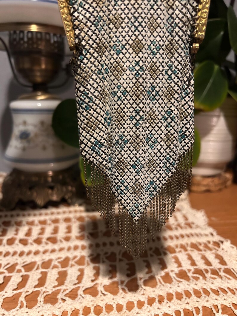 May include: A metal mesh purse featuring a geometric pattern in white, teal, and gold tones. The bag has a gold-toned metal frame at the top and a long, dangling fringe made of thin gold-toned metal chains at the bottom point.
