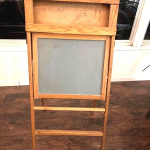 May include: A wooden easel with a gray chalkboard and a light-colored wooden frame. The easel has a rectangular chalkboard and a wooden backboard. The easel is standing on a wooden floor. The easel is designed for art or display purposes.