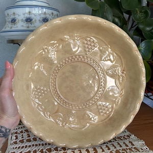 Puede incluir: Un cuenco de cerámica beige con borde festoneado. El cuenco presenta diseños en relieve de uvas y hojas alrededor del borde y un patrón circular central. El cuenco es probablemente vintage y adecuado para servir fruta o ensaladas.