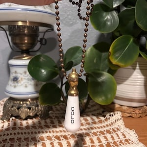 May include: A vintage-style cold water faucet pull chain. The pull features a white, teardrop-shaped ceramic handle with the word "COLD" in black lettering. It has a brass-colored top and is suspended from a matching ball chain.