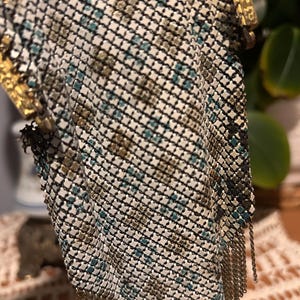 May include: An antique metal mesh handbag with a gold-toned frame and chain fringe. The mesh pattern features a mix of white, black, teal, and brown squares. The bag's clasp and frame are intricately detailed.