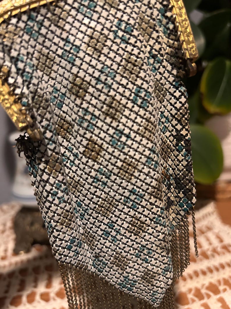May include: An antique metal mesh handbag with a gold-toned frame and chain fringe. The mesh pattern features a mix of white, black, teal, and brown squares. The bag's clasp and frame are intricately detailed.